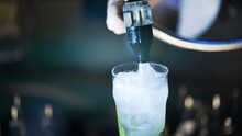 One Mojito Cocktail On Bar Counter, Close Up Shot, Preparing Pouring Soda Into Glass. Cocktail With Mint And Lime On Table In Bar, Blurry Background Of Night Bar Club