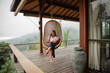 © Yevhenii - Pretty girl sitting on the apartment terrace on the background of mountain landscape and talking the phone, sit on hanging chair