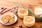 Fototapeta Kuchnia - christmas and seasonal drinks concept - glasses of eggnog with oatmeal cookies, candy canes, sugar, fir tree branches and candle burning on wooden background