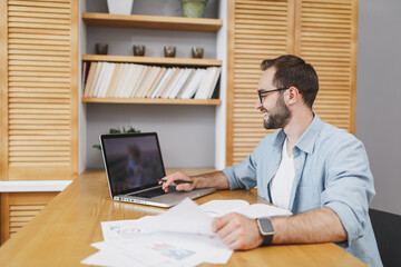 Wall Mural - Handsome smiling attractive successful young bearded business man 20s wearing blue shirt glasses sitting at desk with papers document working on laptop pc computer at home or office.