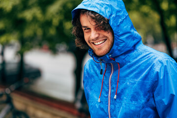 Closeup portrait of a happy man smiling, wearing blue raincoat during the rain outside. Cheerful male enjoying the rain in the city street. The guy has joyful expression in rainy weather.