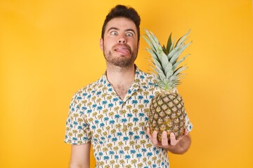 Young man holding pineapple wearing hawaiian shirt over yellow isolated background showing grimace face crossing her eyes and showing tongue Being funny and crazy