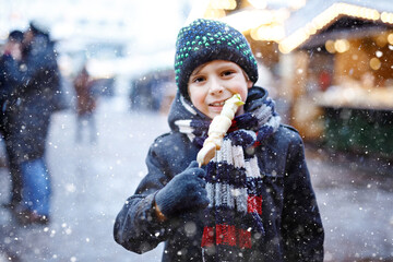 Wall Mural - Little cute kid boy eating white chocolate covered fruits on skewer on traditional German Christmas market. Happy child on traditional family market in Germany during snowy day.