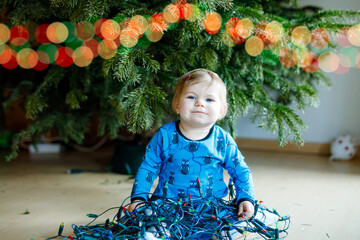 Cute baby girl taking down holiday decorations from Christmas tree. child holding light garland. Family after celebration remove and dispose tree