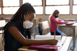 © Wavebreak Media - Mixed Race Girl wearing face mask writing while sitting on her desk at school