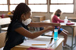 © Wavebreak Media - Girl wearing face mask sanitizing her hands while sitting on her desk at school
