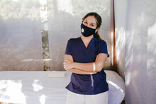 Female Therapist Wearing Mask With Arms Crossed Standing By Massage Table In Spa