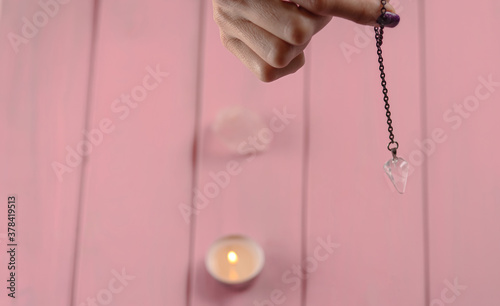 Fotomural Arm of a dowser with hand-held pendulum on a chain with a natural crystal on a background of pink wood texture with burning candles