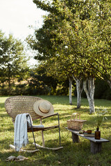 Stylish composition of countryside garden with design rattan armchair, wooden bench, plaid, food, drinks and elegant accessories. A lot of colorful flowers. Summer mood.