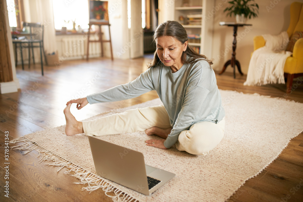 Indoor image of flexible active mature gray haired female sitting on floor in front of open laptop, stretching legs during online yoga lesson, watching live video with professional instructor - obrazy, fototapety, plakaty 
