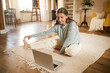 © shurkin_son - Indoor image of flexible active mature gray haired female sitting on floor in front of open laptop, stretching legs during online yoga lesson, watching live video with professional instructor