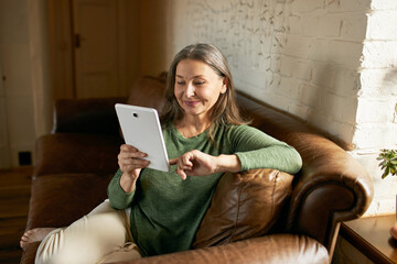 People, communication, electronic gadgets and devices. Beautiful woman pensioner relaxing on comfortable leather sofa with digital tablet, playing video games or chatting online via social networks