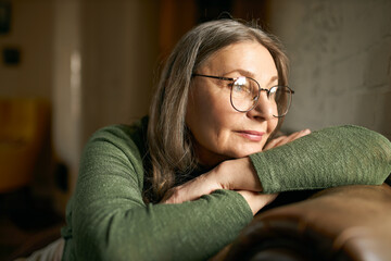 Close up image of beautiful fashionable mature woman wearing stylish round eyeglasses relaxing indoors on leather couch, having pensive deep in thoughts facial expressions, thinking about her life