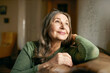 © shurkin_son - Close up image of beautiful positive pensive European female pensioner with gray hair and wrinkled skin relaxing at home on comfortable leather couch, looking up with dreamy thoughtful smile