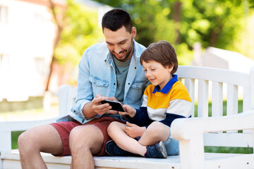 family, fatherhood and technology concept - happy father and little son with smartphone sitting on bench at summer park