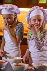 Wall Mural - Little girls in white chef uniform and hats cooking by the table in kitchen. Two cute sisters preparing dough