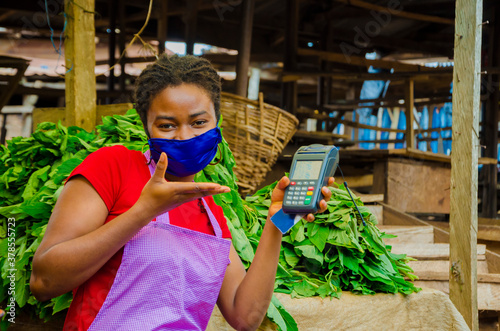 Fotomural a young beautiful african market woman wearing face mask to prevent herself from the society holding a pos machine