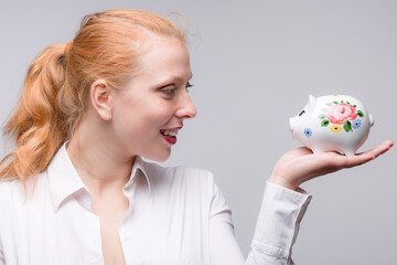 Wall Mural - Young red haired woman looking happy at her piggy bank / porcelain bank