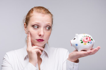 Wall Mural - Young red haired woman looking doubtful to her piggy bank / porcelain bank