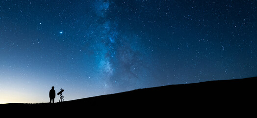 Person observing the blue starry sky with a telescope at night. Silhouette of an astronomer observing the immensity of the universe and the Milky Way