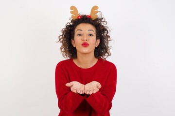 Wall Mural - Young arab woman with curly hair wearing christmas headband on white background sending blow kiss with pout lips and holding palms to send air kiss.