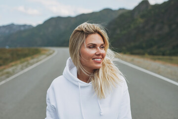 Wall Mural - Beautiful smiling blonde young woman traveler in white hoodie on road, trip to the mountains, Altai