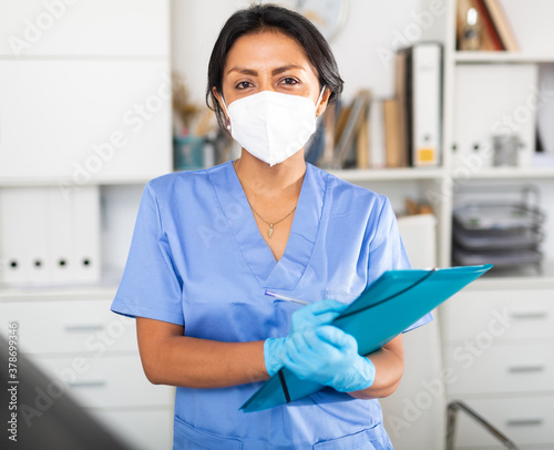 Hispanic woman general practitioner in blue uniform and face mask standing in clinic, filling out clipboard with medical records