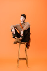 Poster - stylish man in autumn outfit and glasses sitting on wooden stool and looking at camera on orange