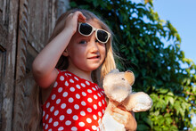 Beautiful Happy Girl In Red Polka Dot Dress With Dog Soft Toy Smiling On Wooden Balcony. Cute Joyful Child With Long Blonde Hair In Sunglasses On Green Natural Wild Grape Backdrop. Street Portrait.