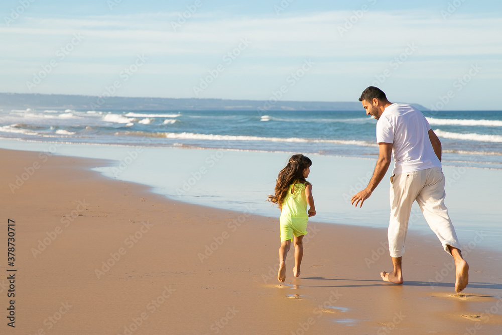 Father and little daughter running on wet golden sand on beach. Girl enjoying leisure time with dad by sea. Back view. Family outdoor activities concept