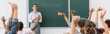 Back View Of Multicultural Pupils With Hands In Air, And Teacher Standing At Chalkboard With Back To School Inscription, Horizontal Image