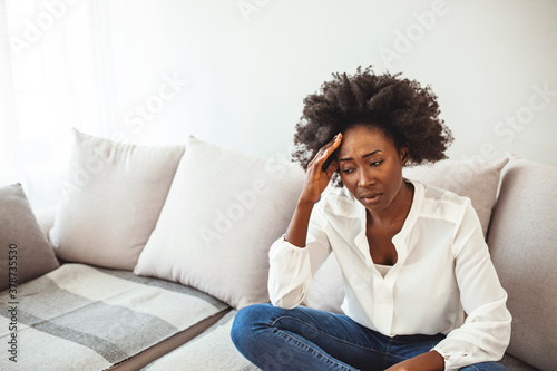 African woman sitting on couch feels unhappy having problems. Need to take some pills. Cannot stand this pain. Frustrated lady feeling severe headache