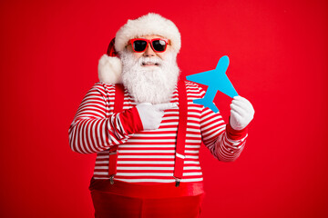 Poster - Photo of old man grey beard hold direct finger paper plane smile wear santa claus x-mas costume suspenders sunglass striped shirt cap isolated red color background