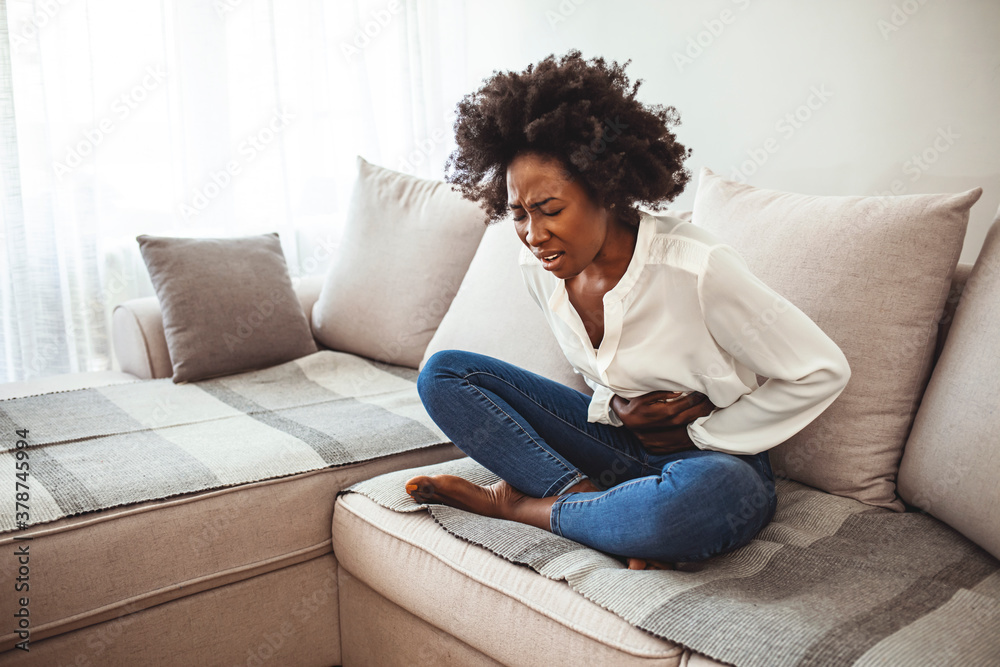 View of young woman suffering from stomachache on sofa at home. Woman sitting on bed and having stomach ache. Young woman suffering from abdominal pain while sitting on sofa at home - obrazy, fototapety, plakaty 