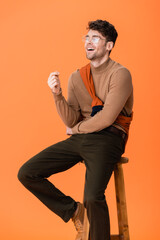 Poster - excited man in autumn outfit and glasses sitting on wooden stool and laughing on orange