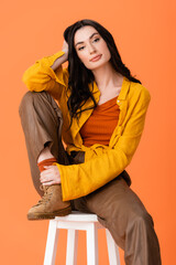 Wall Mural - trendy woman in autumn outfit sitting on white stool and looking at camera isolated on orange