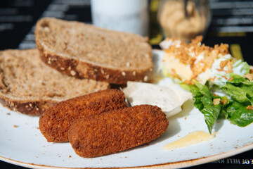 Two delicious Dutch croquettes on a brown bun with mustard, standing on a terrace table, people eating and drinking in the background