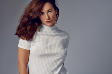 Sexy attractive young woman with shoulder length auburn hair wearing a high necked white summer top giving the camera a sultry sensual look over a grey background with copy space