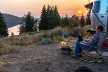 Person Sitting On A Rocking Chair By The Side Of A Travel Trailer, RV, Campfire Working On A Laptop, PC As The Sun Setting Over A Distance Hill, Mountain, Half Moon Lake, Pinedale, Wyoming