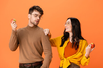 Poster - stylish couple in glasses and autumn outfit holding apples while looking at each other isolated on orange