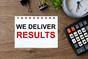 Poster - We Deliver ResultsWE DELIVER RESULTS. text on white paper on wood background near calculator, paper clips, alarm clock, PLANTS