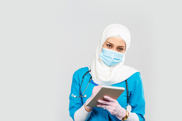 Wall Mural - friendly muslim doctor or nurse wearing hijab and medical face mask with stethoscope writing prescription to patient on gray background