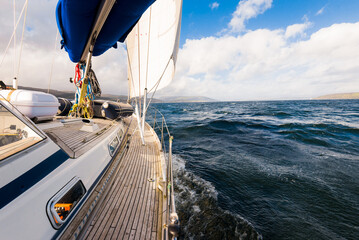 Wall Mural - Sloop rigged modern yacht with wooden teak deck sailing near the rocky shore of the Firth of Clyde river. Dramatic sky. Scotland, UK. Travel destinations, eco tourism, vacations, pure nature