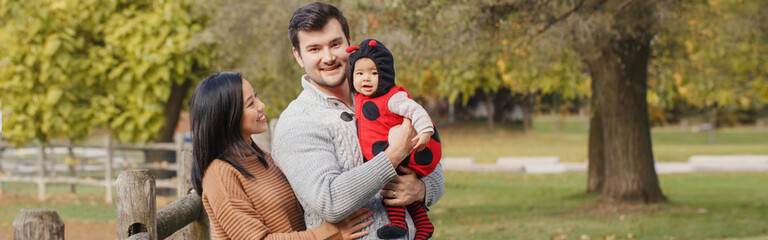 Wall Mural - Happy smiling Asian Chinese mother and Caucasian father dad with baby girl in ladybug costume. Family in autumn fall park outdoors. Halloween or Thanksgiving holiday. Web banner header.