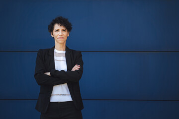 Portrait of confident mature female entrepreneur standing with arms crossed against blue wall
