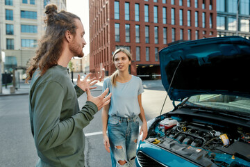 Wall Mural - Portrait of young man talking, explaining the failure to attractive woman while they both standing near the broken car with open hood on the street