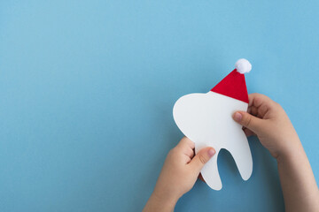 Little child holding cute decorative tooth in a red Santa Claus hat on light blue background. Christmas and New Year. Creative medical winter postcard for dentistry. Top view, copy space, flat lay