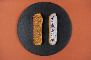 Wall Mural - Raspberry and caramel eclair on round black rock stand on brown background. Two eclairs with glaze in cafe. View from above. Cake with choux cream on slate stand. Tasty dessert concept.