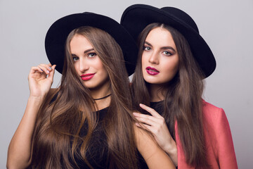 Close up beauty  fashion image of two girls, sisters, posing on grey background, hugging, smiling. Wearing stylish pink  coat and black hat, short top and evening dress.