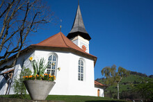 White Church In The French Speaking Part Of Switzerland In The Middle Of A Little Country Side Town With A Beautiful Flower Vase In Front Of The Scene